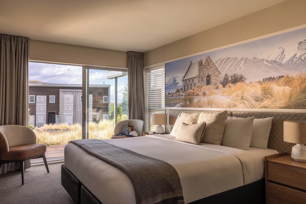 Habitación de hotel con una cama grande, una silla, una mesita de noche y ventana con vistas a un edificio y montañas.