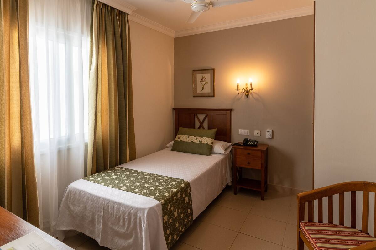 A hotel room with a single bed, a bedside table with a phone, a wall-mounted light, and a chair.