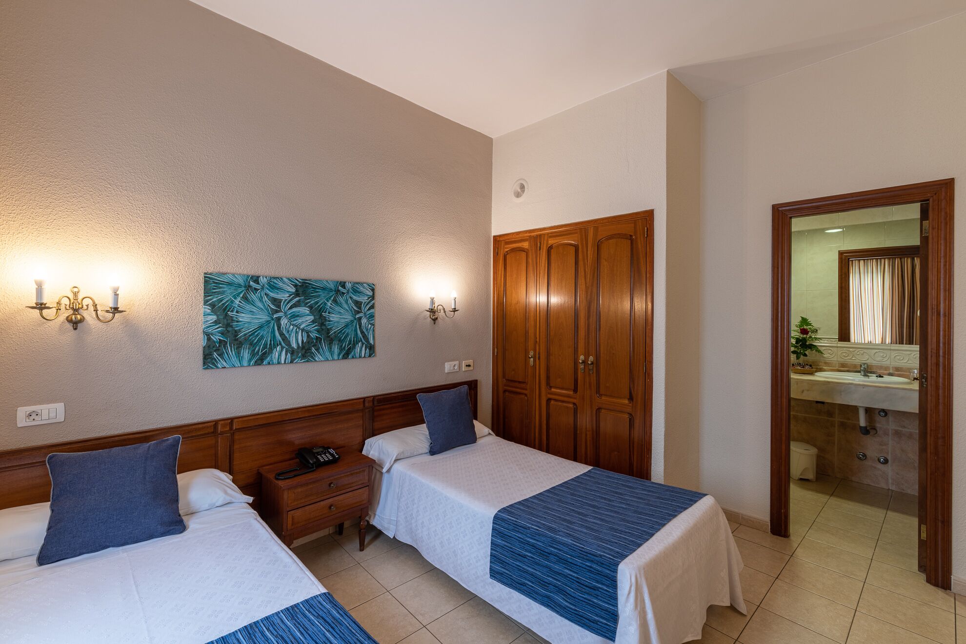 A hotel room with two beds, a wooden wardrobe, a bathroom with a mirror and sink, and a wall-mounted artwork.