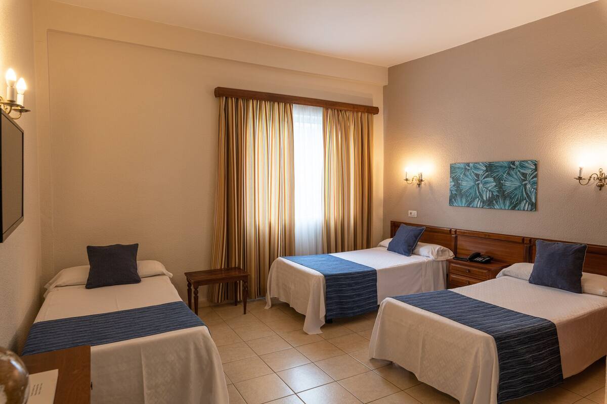 A hotel room with two beds, a small table, a nightstand, a painting on the wall, and a window with curtains.