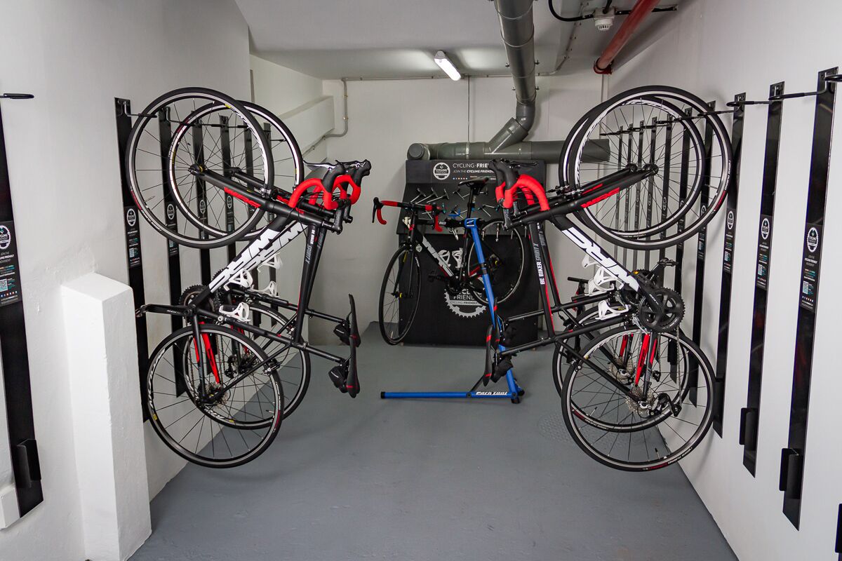Zona de almacenamiento para bicicletas con dos bicicletas aseguradas en soportes, un perchero azul en el centro y un soporte de bicicleta montado en la pared a la derecha.