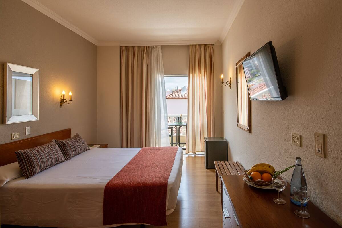A hotel room with a bed, a television mounted on the wall, a small table with a fruit bowl and glasses, and a view of the outside through the window.