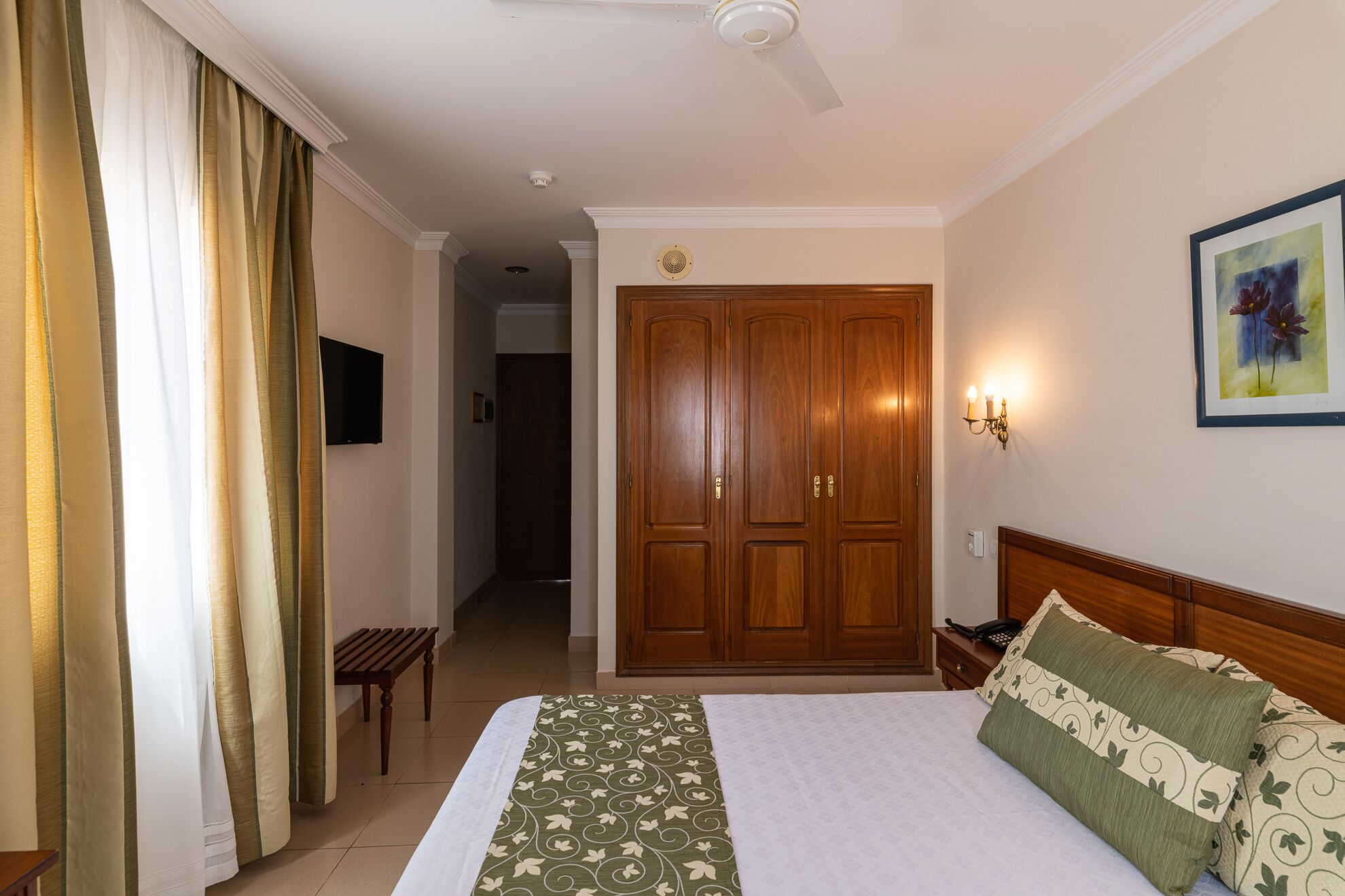 A hotel room with a bed, a wooden wardrobe, a bedside table, a lamp, and a painting on the wall.