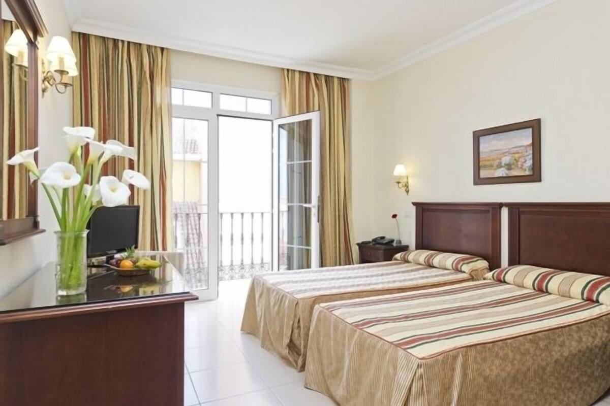 A hotel room with a bed, a desk with a vase of flowers, a painting on the wall, and a view through a sliding door.