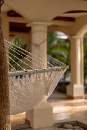 A hammock suspended between two columns in a covered outdoor area with a tiled floor and palm trees in the background. - RipJack Inn (Cabo Velas)