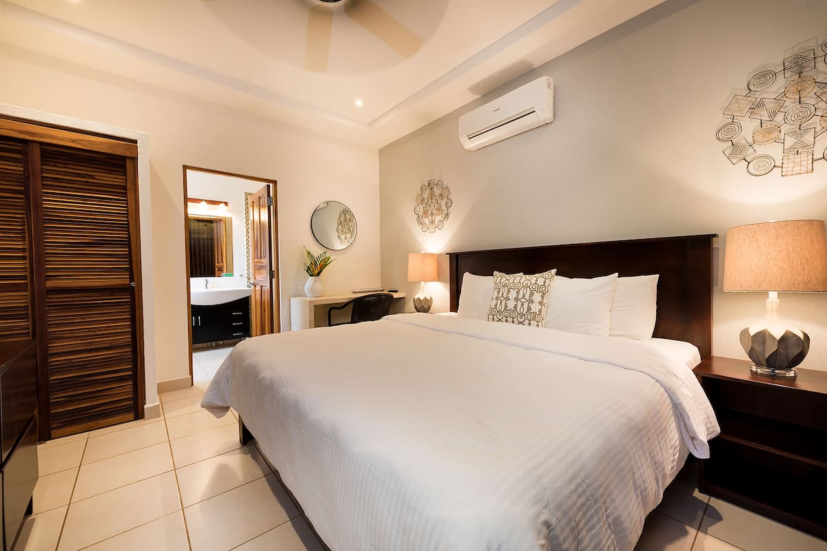 Una habitación de hotel con una cama grande, una mesita de noche con lámpara, aire acondicionado instalado en la pared y un espejo.