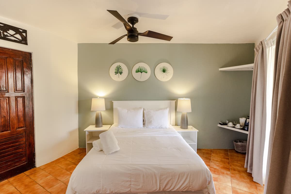 Habitación de hotel ordenada con cama, mesitas de noche, lámparas, ventilador de techo y obras de arte colgadas en la pared.