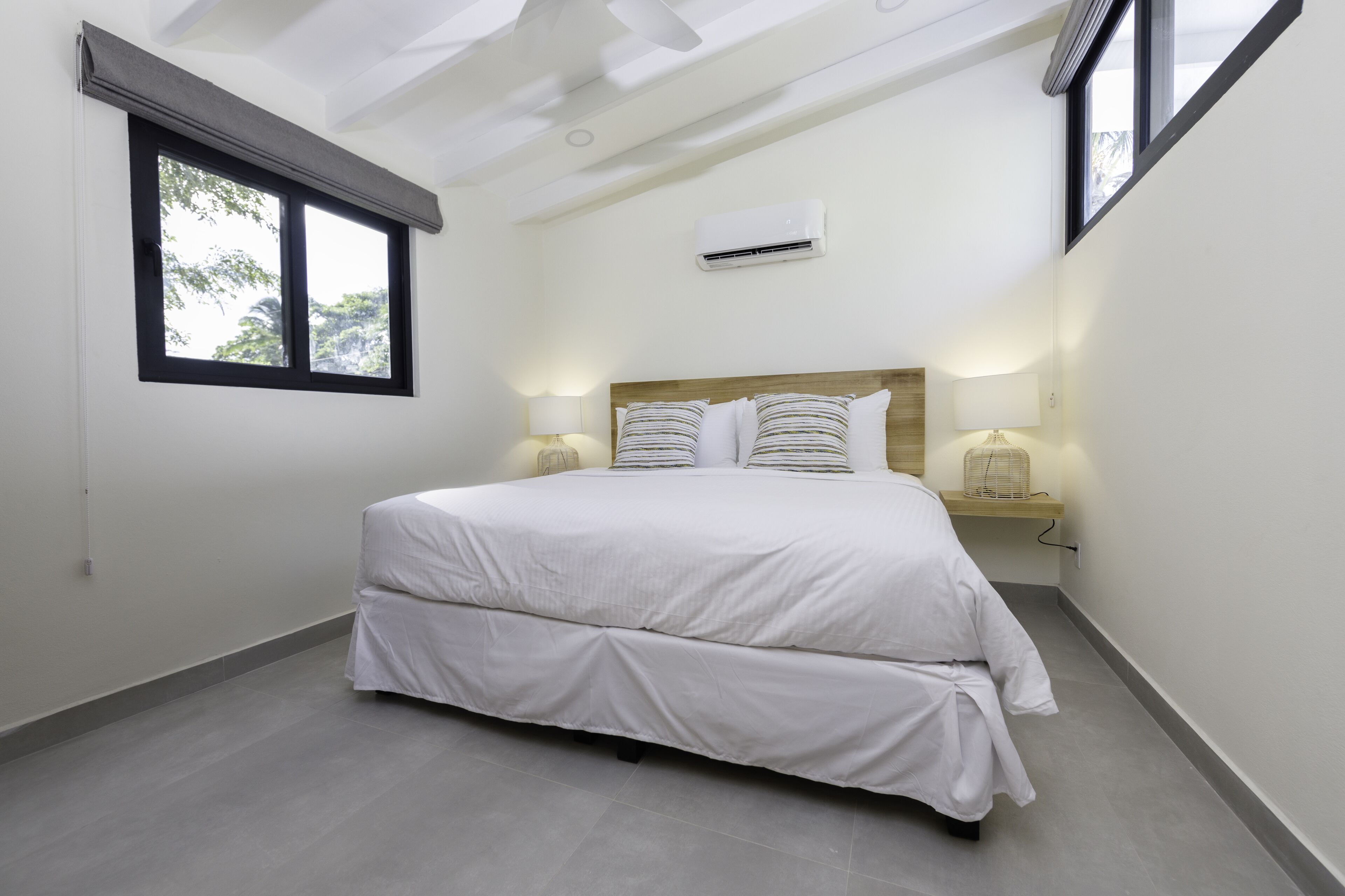 A bedroom with a large bed, two bedside lamps, a window with blinds, and an air conditioning unit.