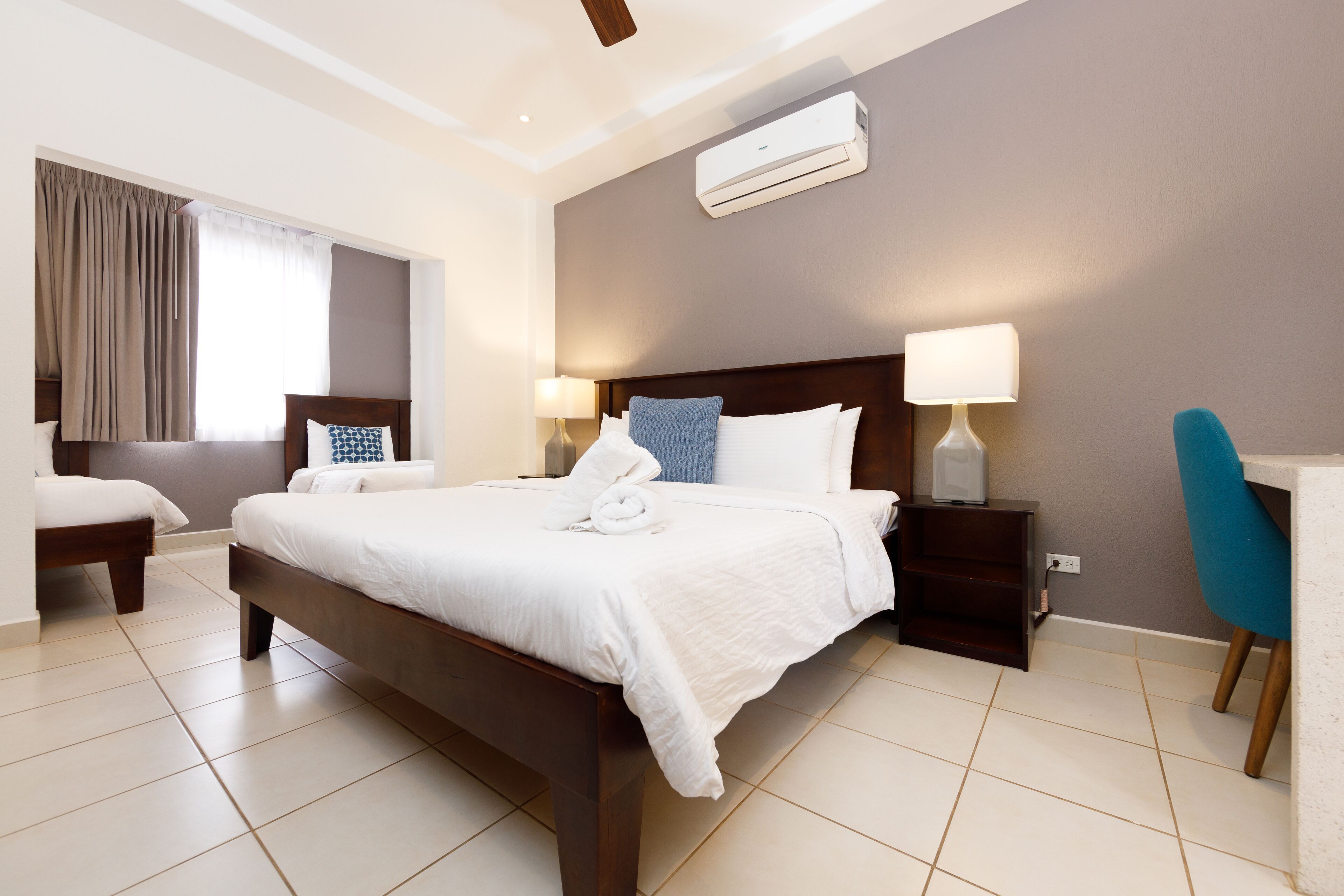 Quarto de hotel com uma cama grande, duas mesas de cabeceira com luminárias, uma escrivaninha com cadeira e um aparelho de ar condicionado na parede.