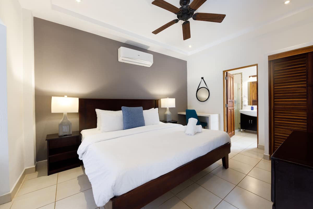 Habitación de hotel con una cama grande, dos mesitas de noche con lámparas, un ventilador de techo, un equipo de aire acondicionado y un baño visible a través de una puerta abierta.