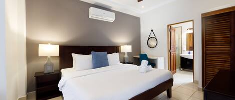 A hotel room with a large bed, two bedside tables with lamps, a ceiling fan, an air conditioning unit, and a bathroom visible through an open door.