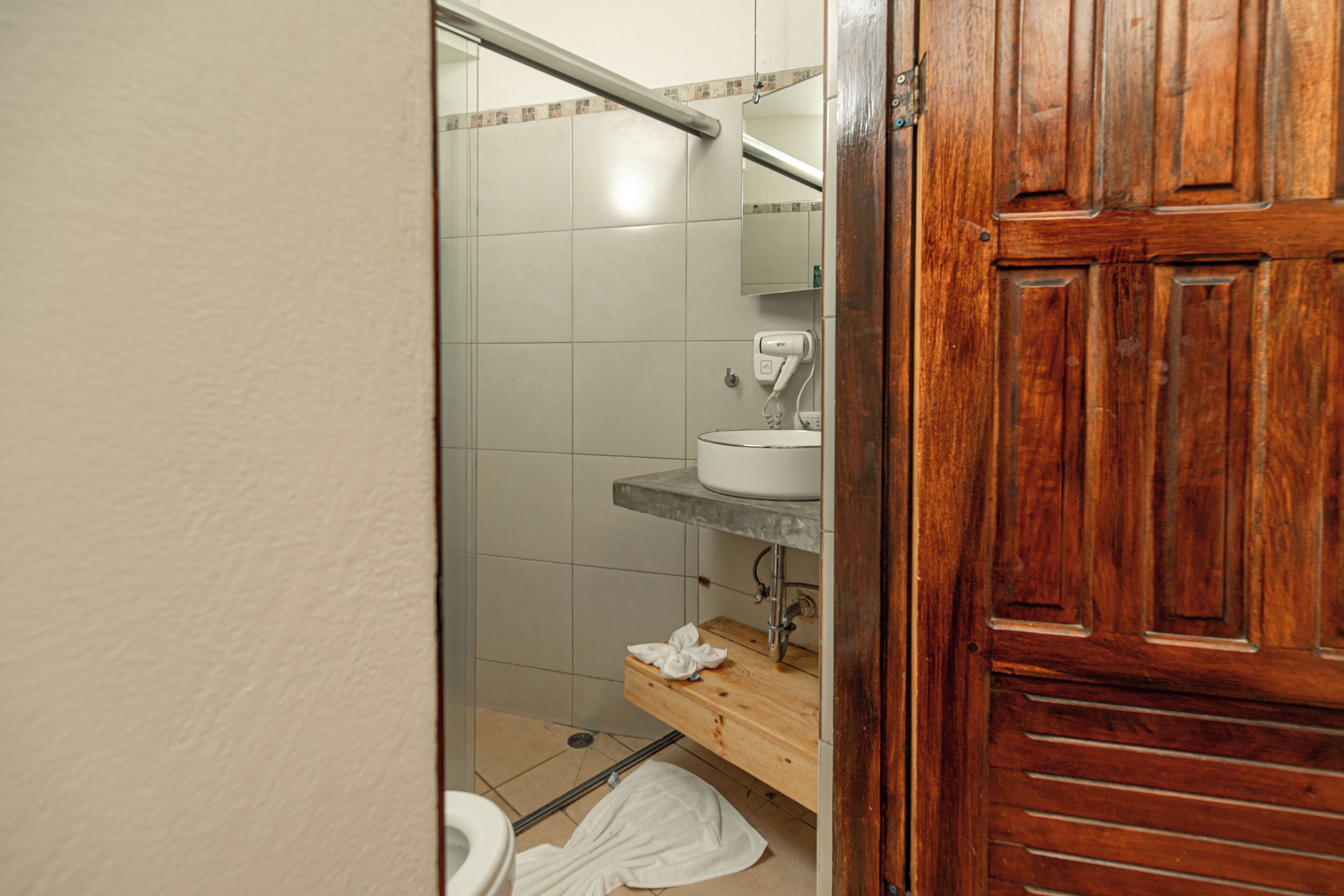 Un baño con puerta de madera, lavamanos blanco, espejo y zona de ducha.