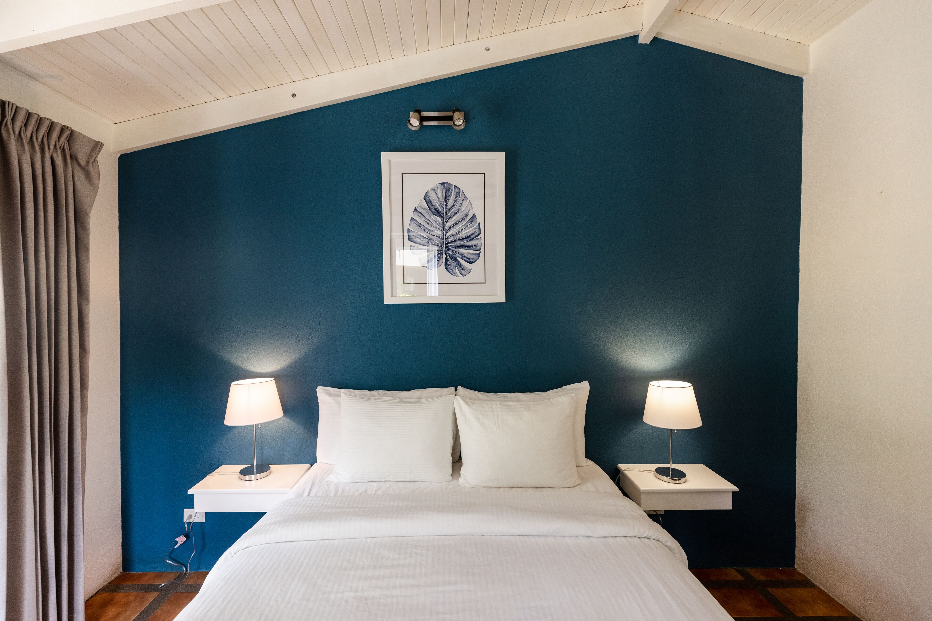 Un dormitorio con una cama, dos mesitas de noche con lámparas, una obra de arte enmarcada en la pared y una ventana con cortinas.