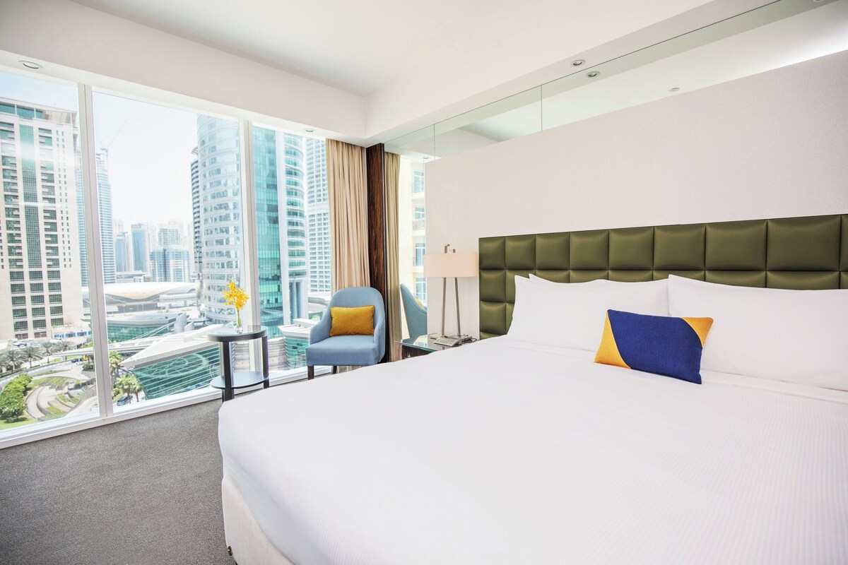 Ein Hotelzimmer mit einem großen Bett, Blick auf die Stadt und einem Sessel am Fenster.