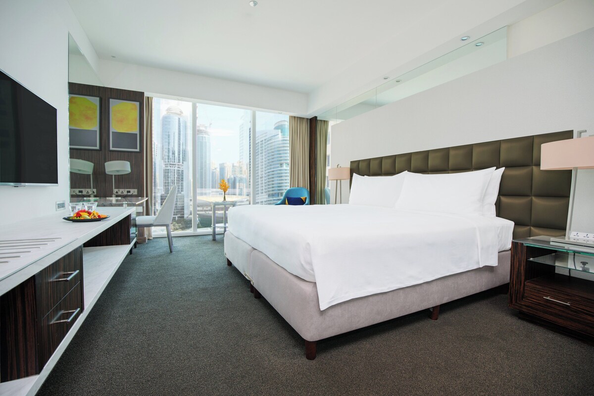 Ein modernes Hotelzimmer mit einem großen Bett, einem Schreibtisch mit Stuhl, einem Fernseher und Blick auf die Skyline der Stadt.