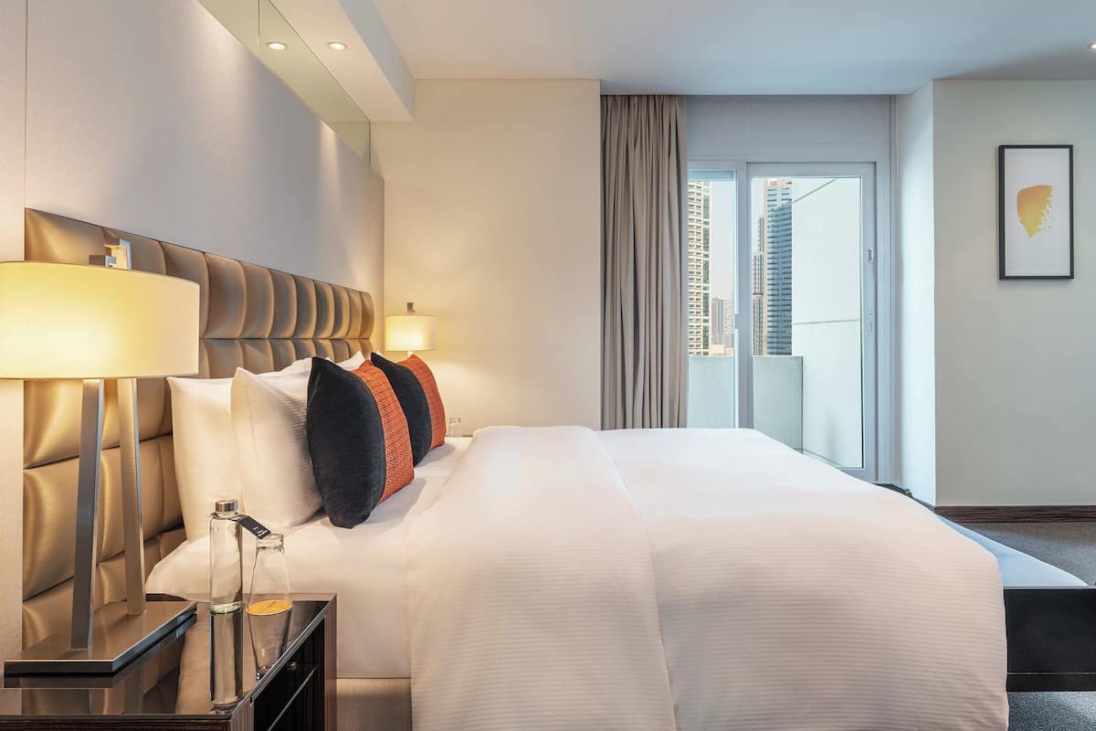 Ein modernes Hotelzimmer mit einem großen Bett, einem Nachttisch mit Lampe und Blick auf die Stadt durch das Fenster.