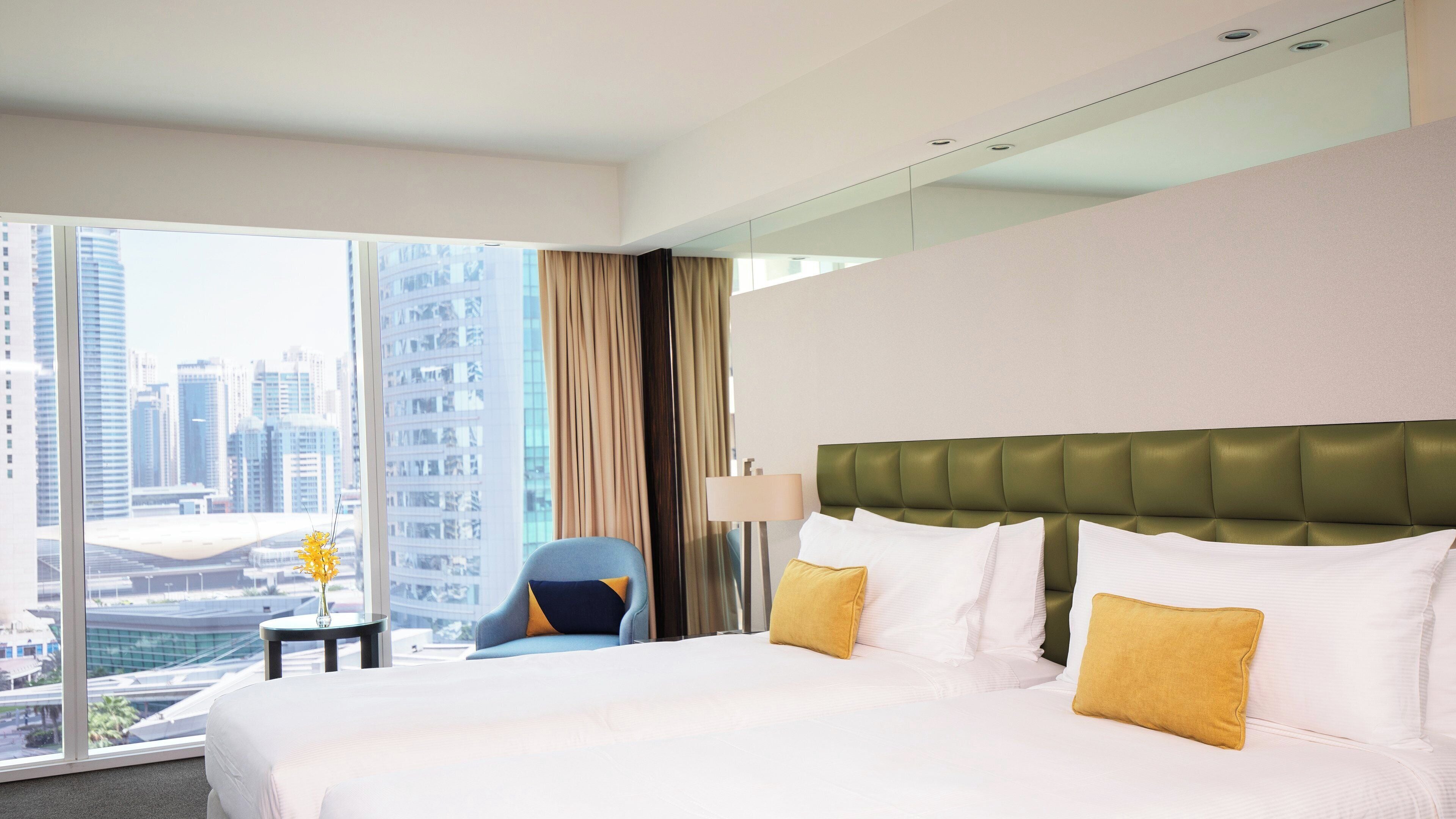 A hotel room with a large bed, a chair, a lamp, and a view of a cityscape.