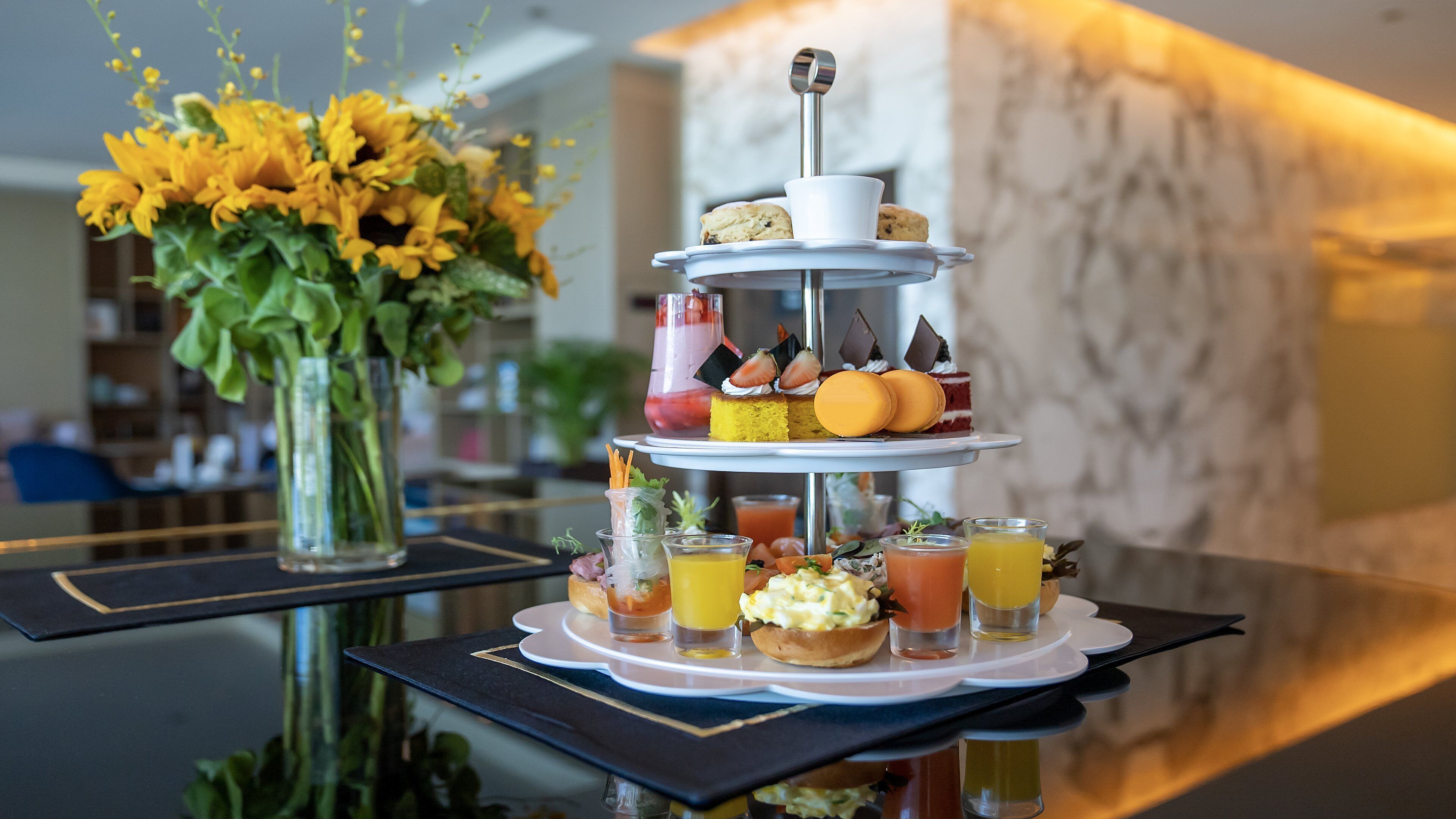 Ett dessertbord i tre våningar med olika sorters bakverk och drycker, placerat på ett bord med reflekterande yta i en hotelllobby.