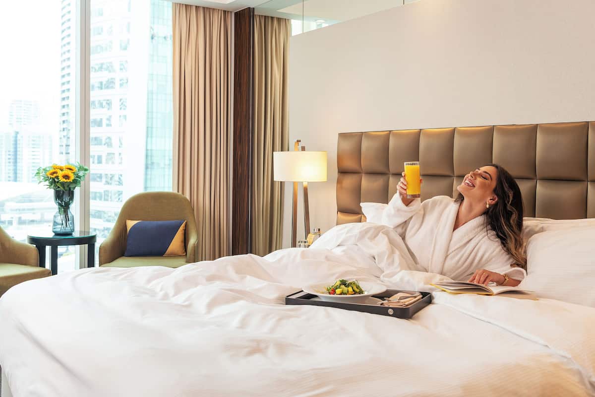 Ein Hotelzimmer mit Bett, Nachttisch mit Lampe, Stuhl und Blick auf die Stadt.