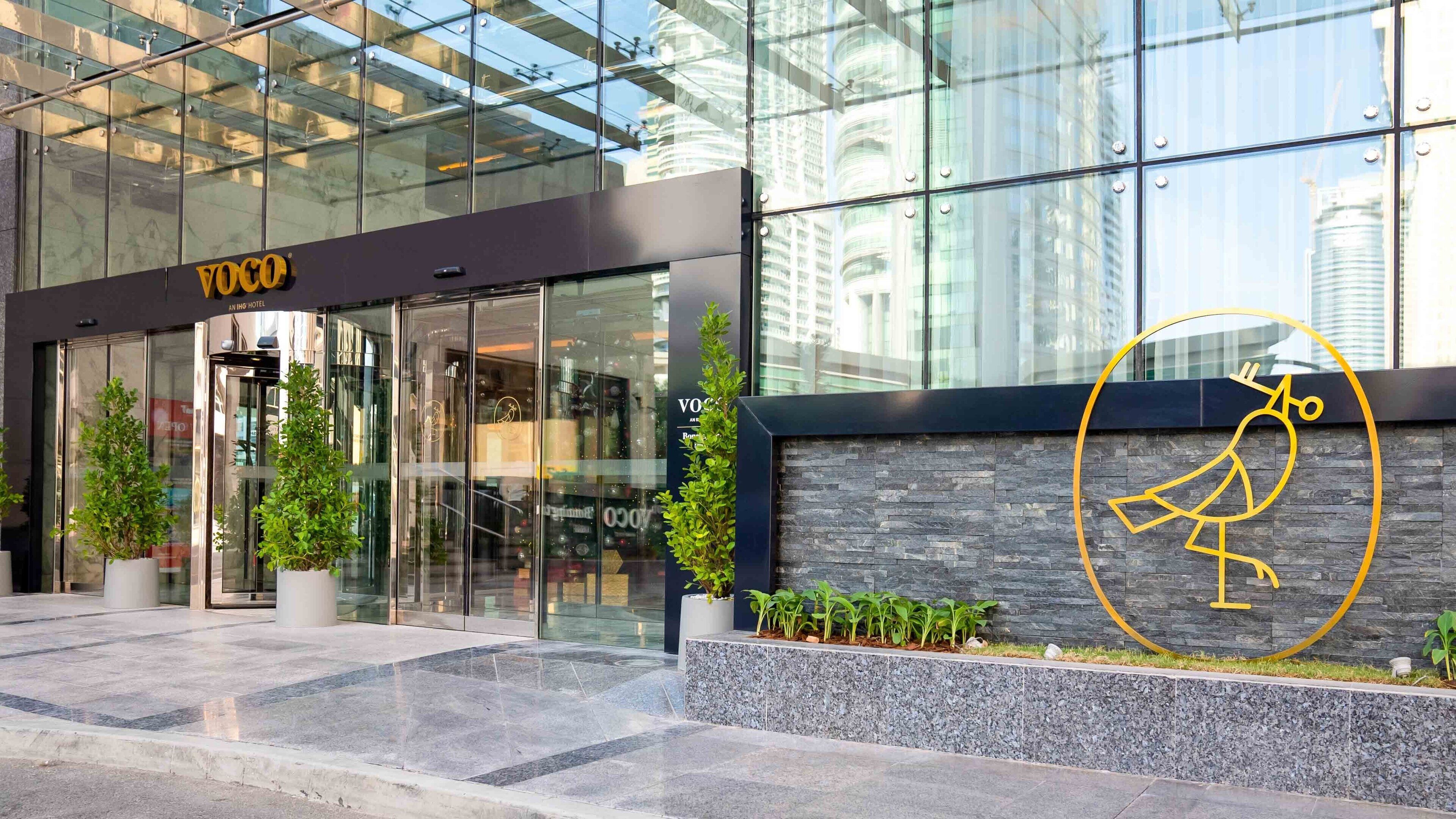 A modern hotel entrance with glass doors, a logo, and a landscaped area with plants.