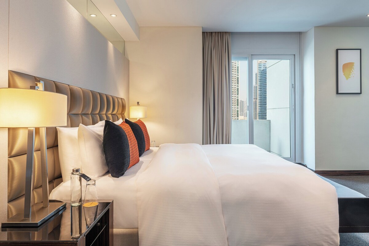 Ein modernes Hotelzimmer mit einem großen Bett, einem Nachttisch mit Lampe und Blick auf die Stadt durch das Fenster.