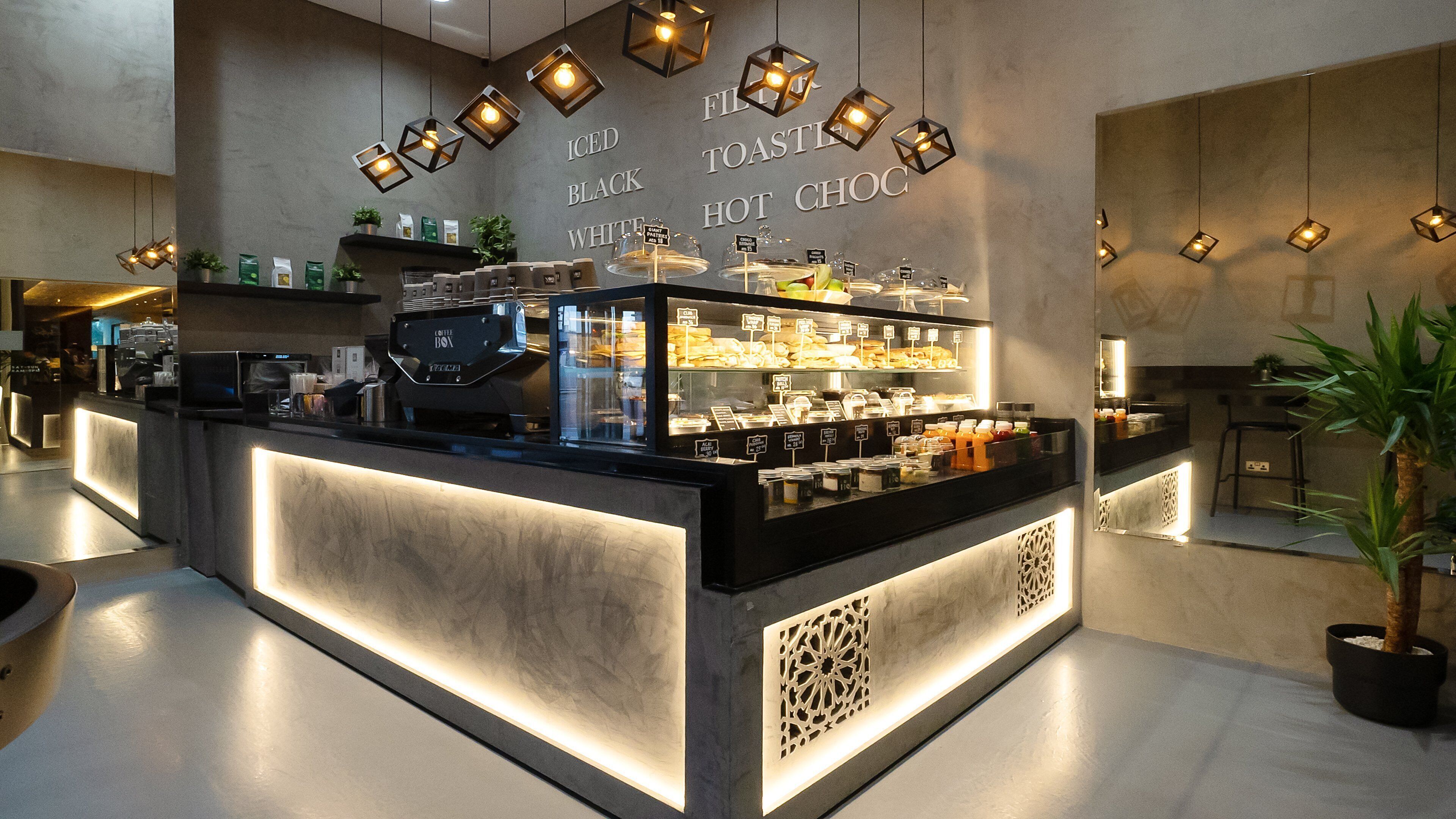 A modern coffee shop with a counter displaying various coffee beans and pastries, a coffee machine, and hanging lights.