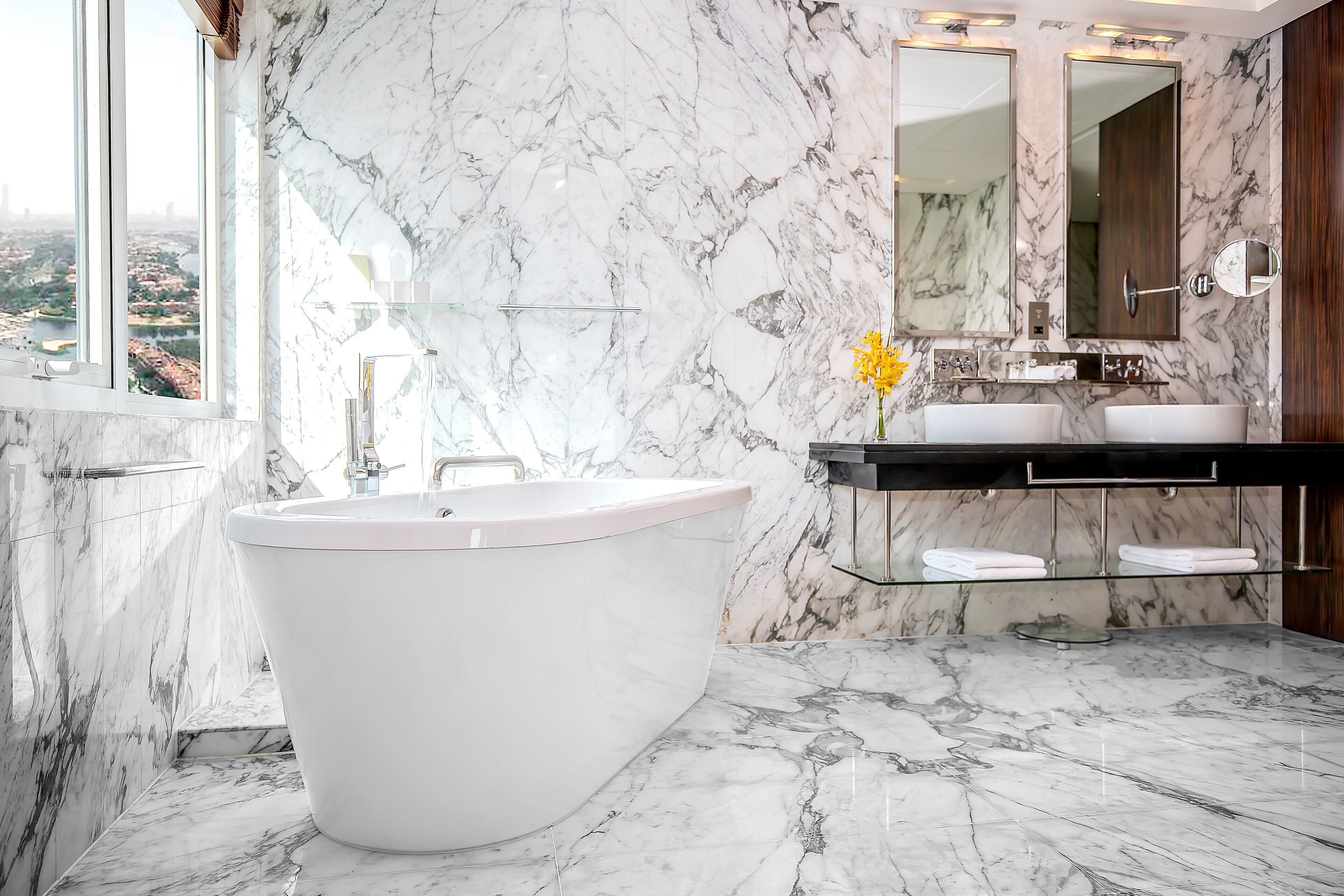 A modern bathroom with a freestanding bathtub, marble walls, a large mirror, and a view of the city.