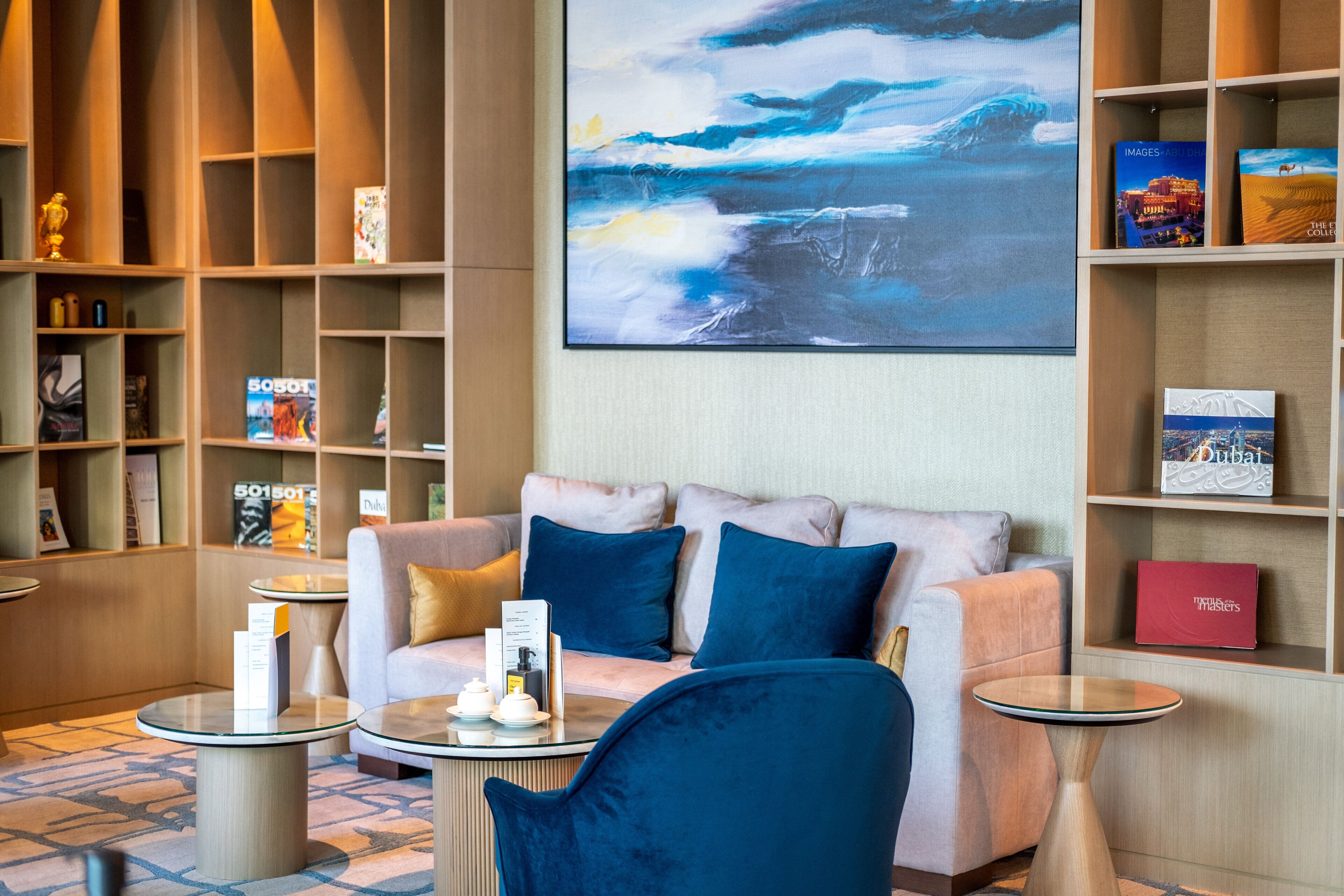 Un hall d’hôtel moderne avec un canapé, deux tables d’appoint, une grande peinture abstraite et des étagères remplies de livres et d’objets décoratifs.