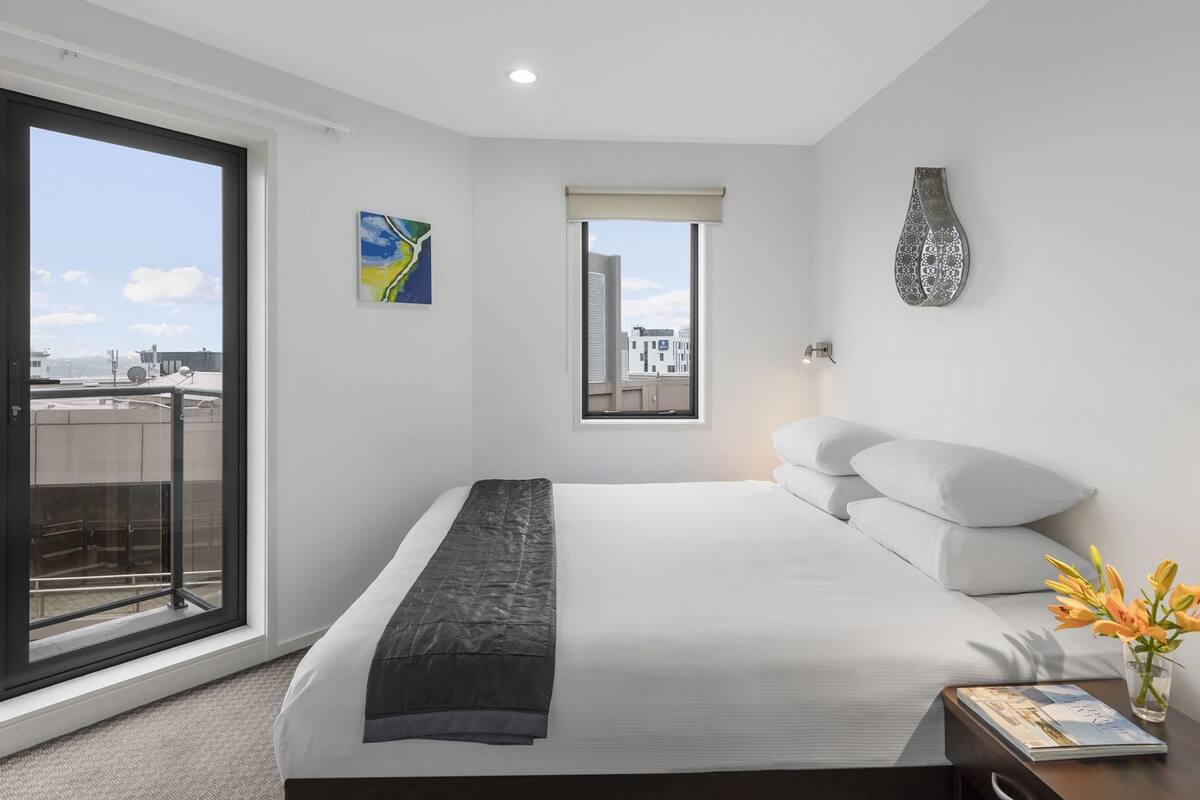 A modern hotel room with a large bed, a window with a city view, a decorative wall piece, and a small table with a vase.