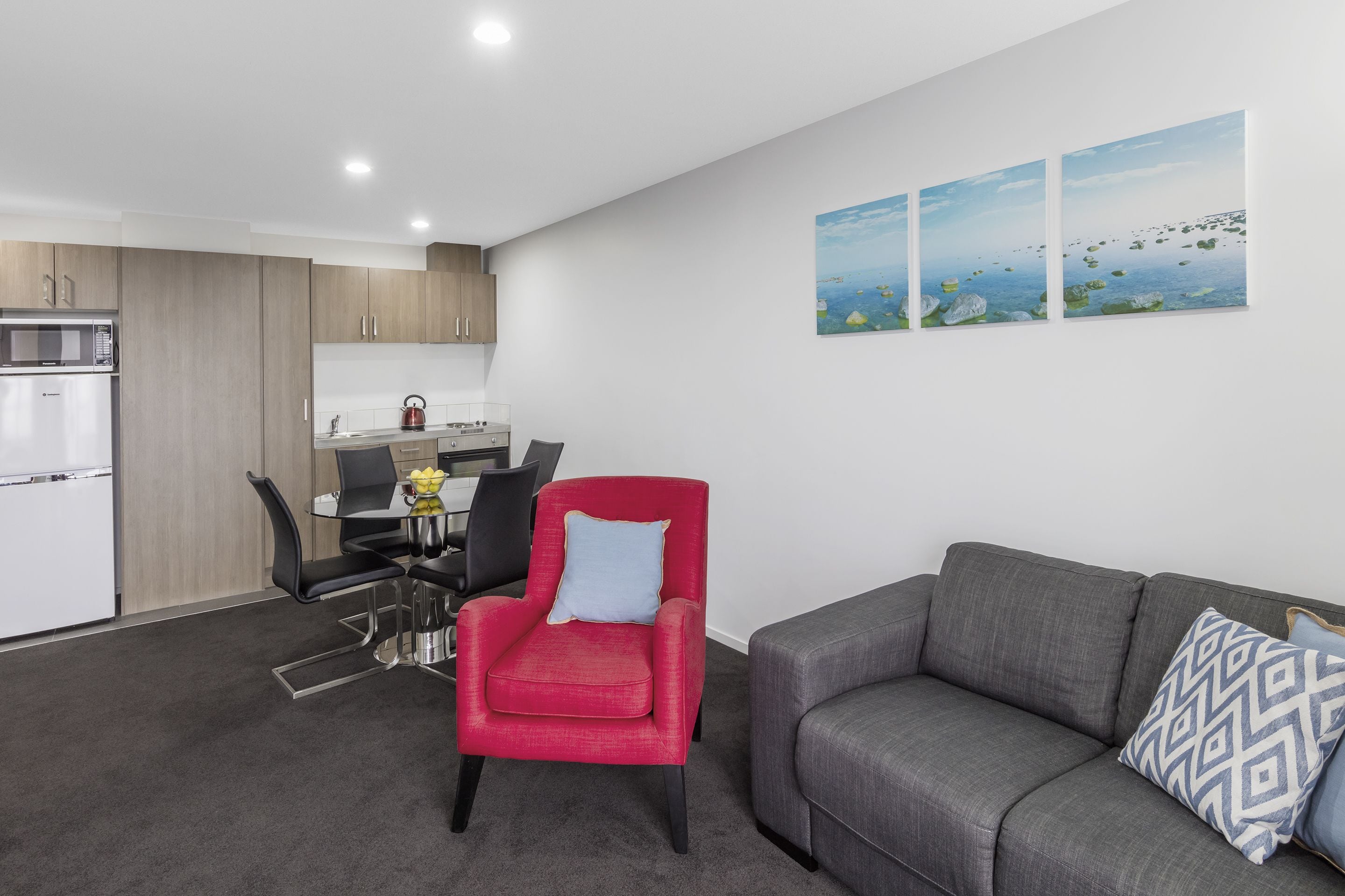 Een moderne woonkamer met een grijze bank en een rode fauteuil, een eethoek met een glazen tafel, en een keuken met houten kastjes en een witte koelkast.