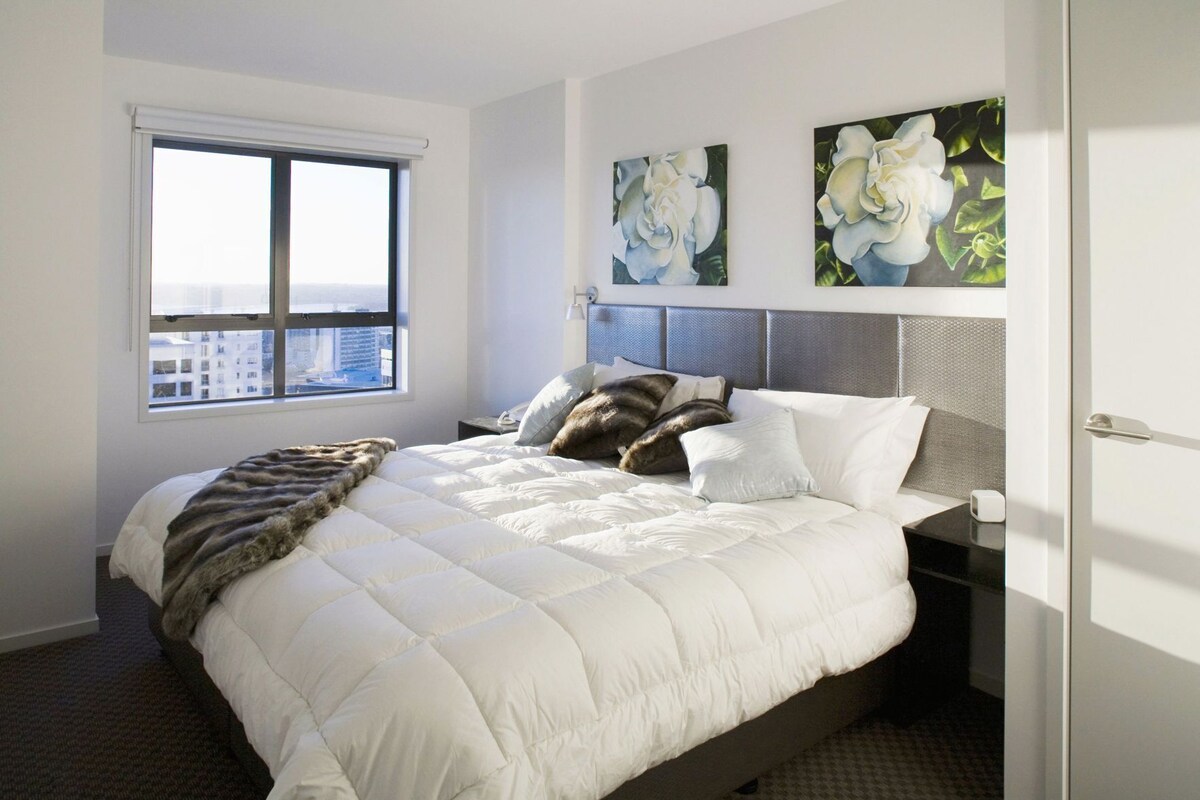 A hotel room with a large bed, a window with a city view, and two floral paintings on the wall.