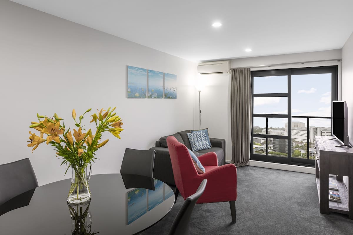 A modern hotel room with a large window, a red armchair, a glass coffee table, and a vase of flowers.