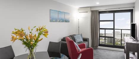 Une chambre d’hôtel moderne avec une grande fenêtre, un fauteuil rouge, une table basse en verre et un vase de fleurs.