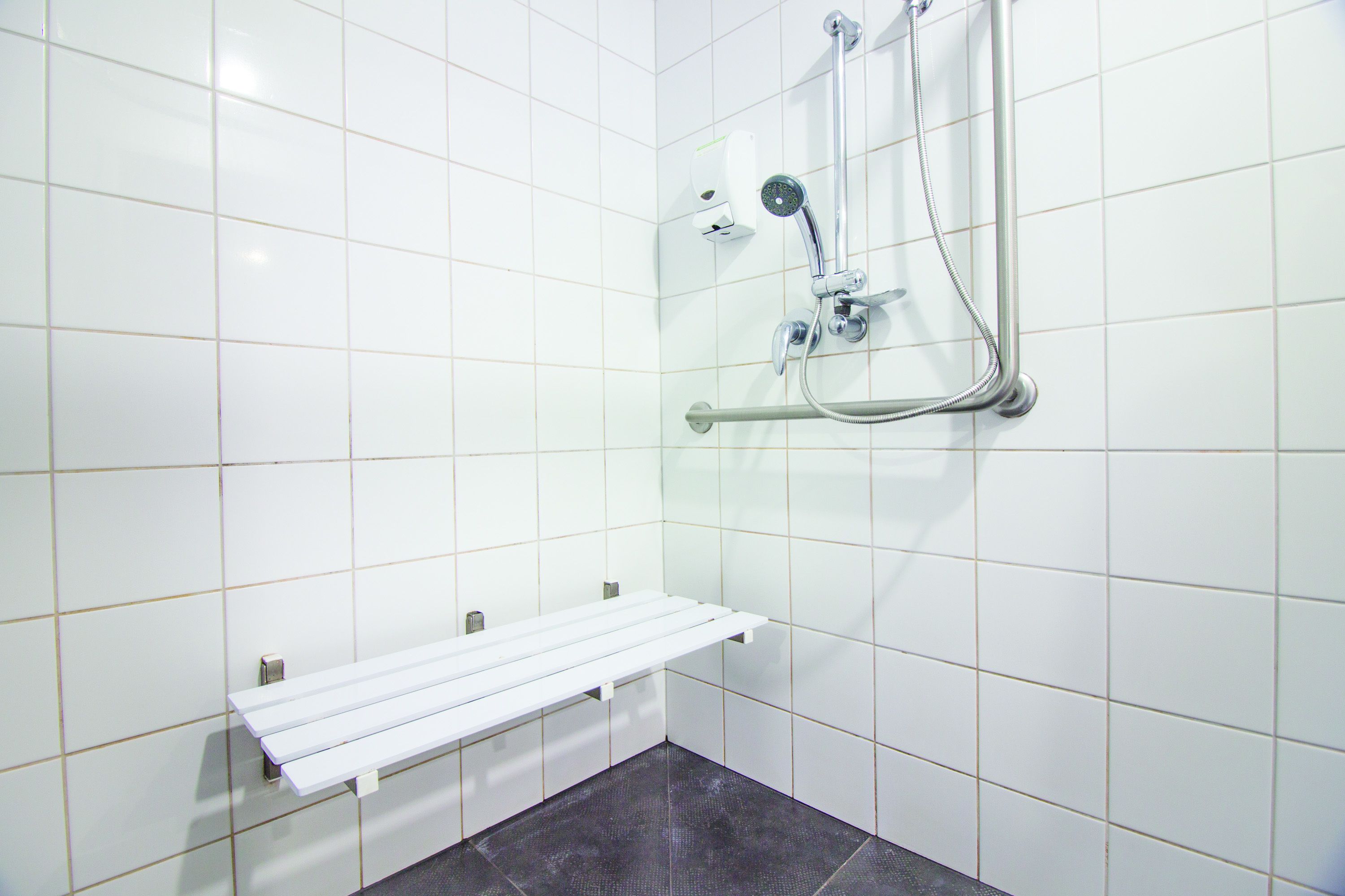 A bathroom with white tiled walls, a shower with a handheld showerhead, and a white bench.
