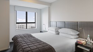 Habitación de hotel moderna con una cama grande, una mesita de noche con libros y una botella, una ventana con vistas a la ciudad y un cabecero con diseño texturizado.