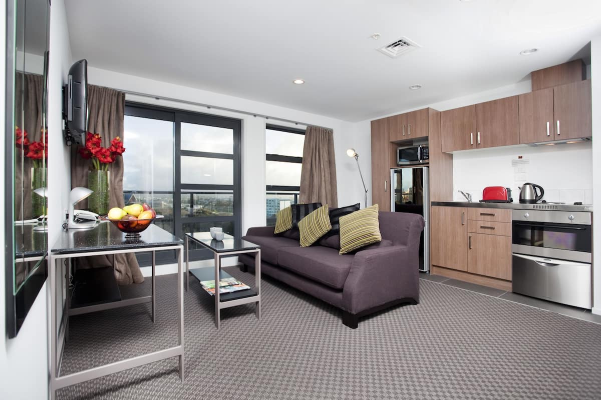 A modern living room with a sofa, coffee table, and a TV mounted on the wall. There is a kitchen area with cabinets and appliances.