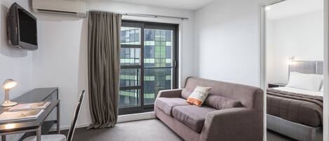 A modern hotel room with a sofa, a desk with a lamp, a flat-screen TV, and a large window with curtains.