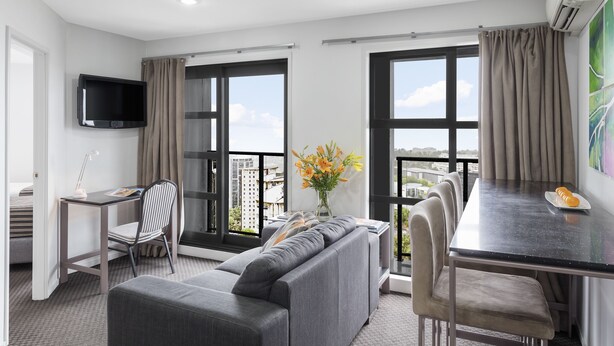 Una habitación de hotel moderna con sofá, mesa de comedor y sillas, televisor en la pared y ventanas grandes con cortinas.