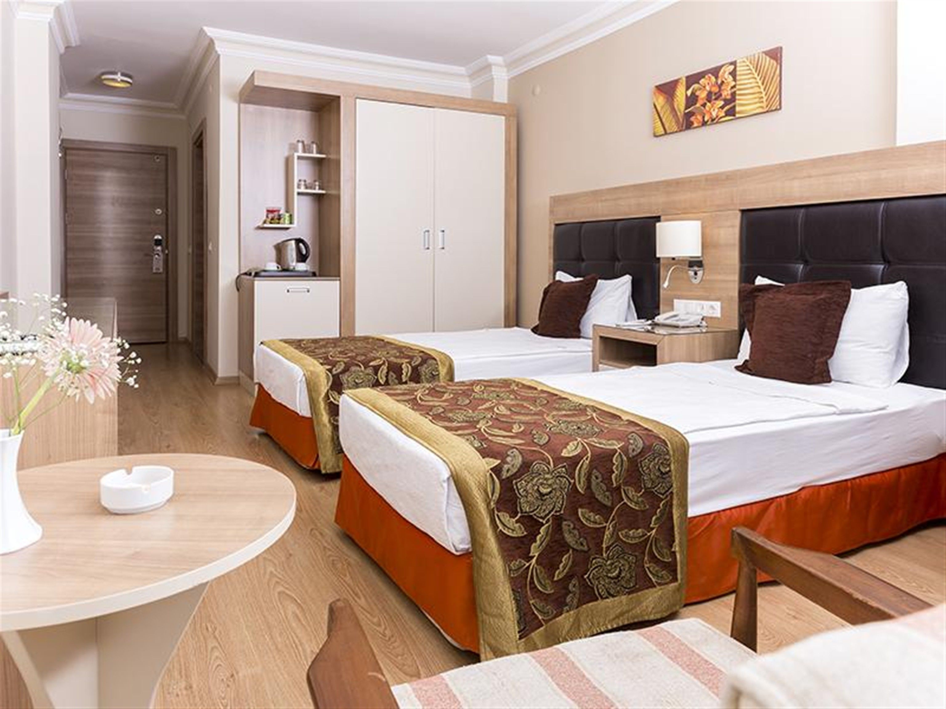 A hotel room with two beds, a desk, a chair, a small table, a vase with flowers, and a painting on the wall.