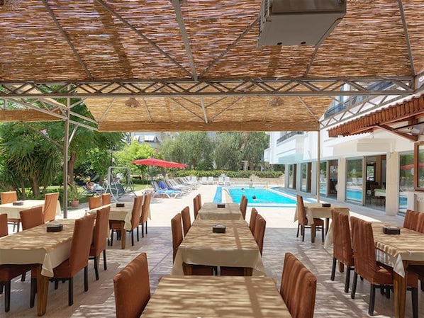 An outdoor dining area with tables and chairs, a pool in the background, and a covered walkway.