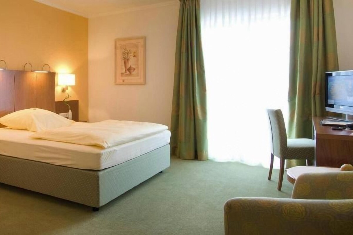 Une chambre d’hôtel comprenant un lit, une table de chevet, une lampe, une chaise, un bureau, une télévision et des rideaux.