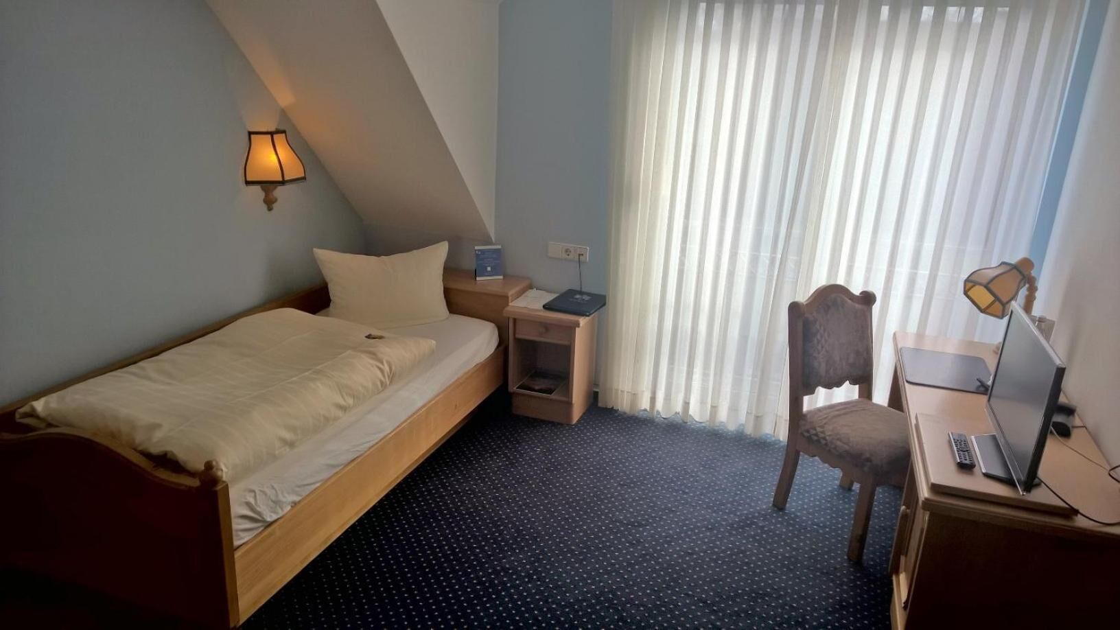 Ein kleines, ordentlich eingerichtetes Hotelzimmer mit einem Einzelbett, einem Schreibtisch mit einem Laptop, einem Stuhl, einem Nachttisch und einem Fenster mit Vorhängen.