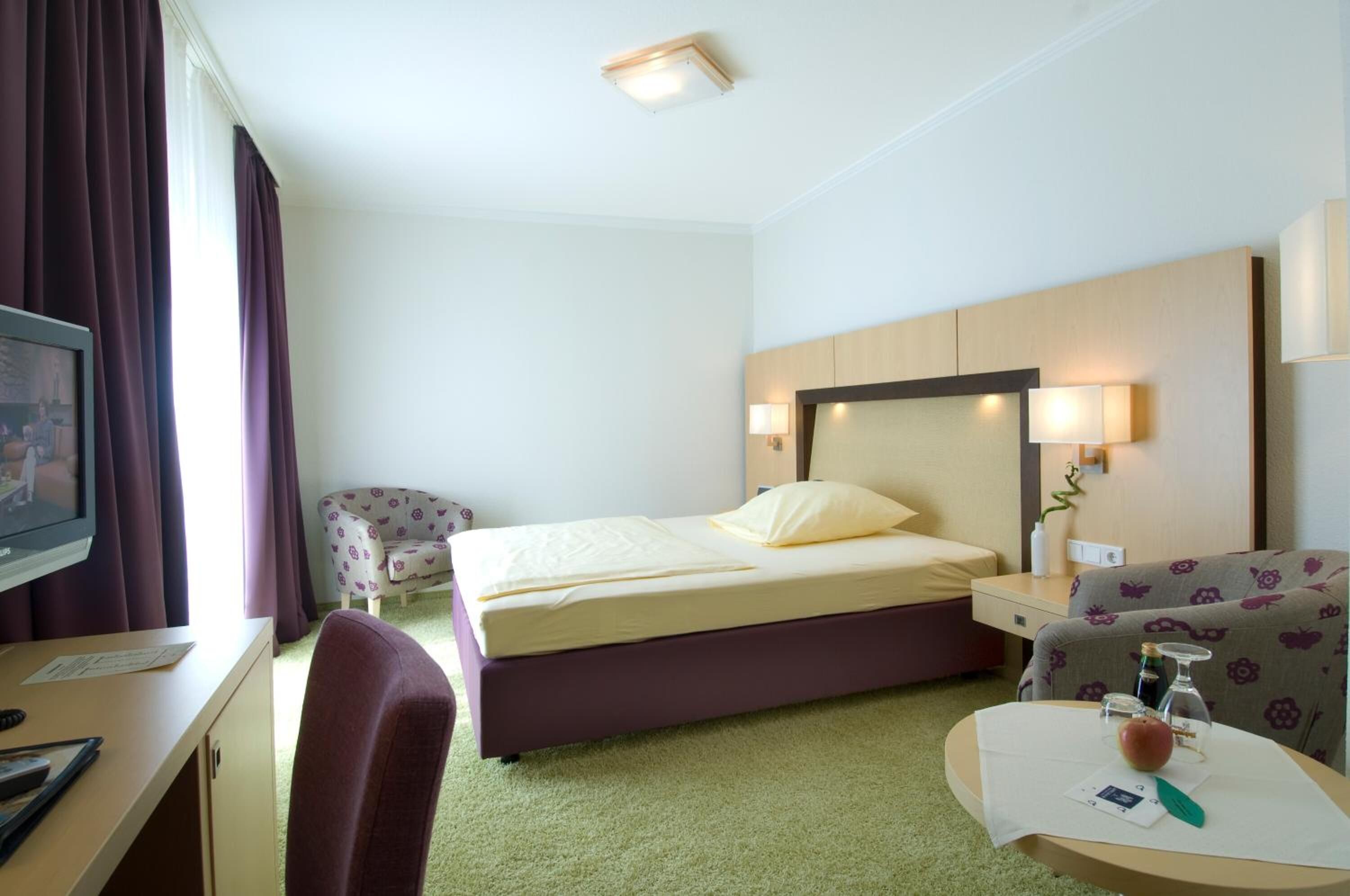 A hotel room with a bed, a desk with a computer, a chair, a small table with a vase and a book, and a wall-mounted TV.