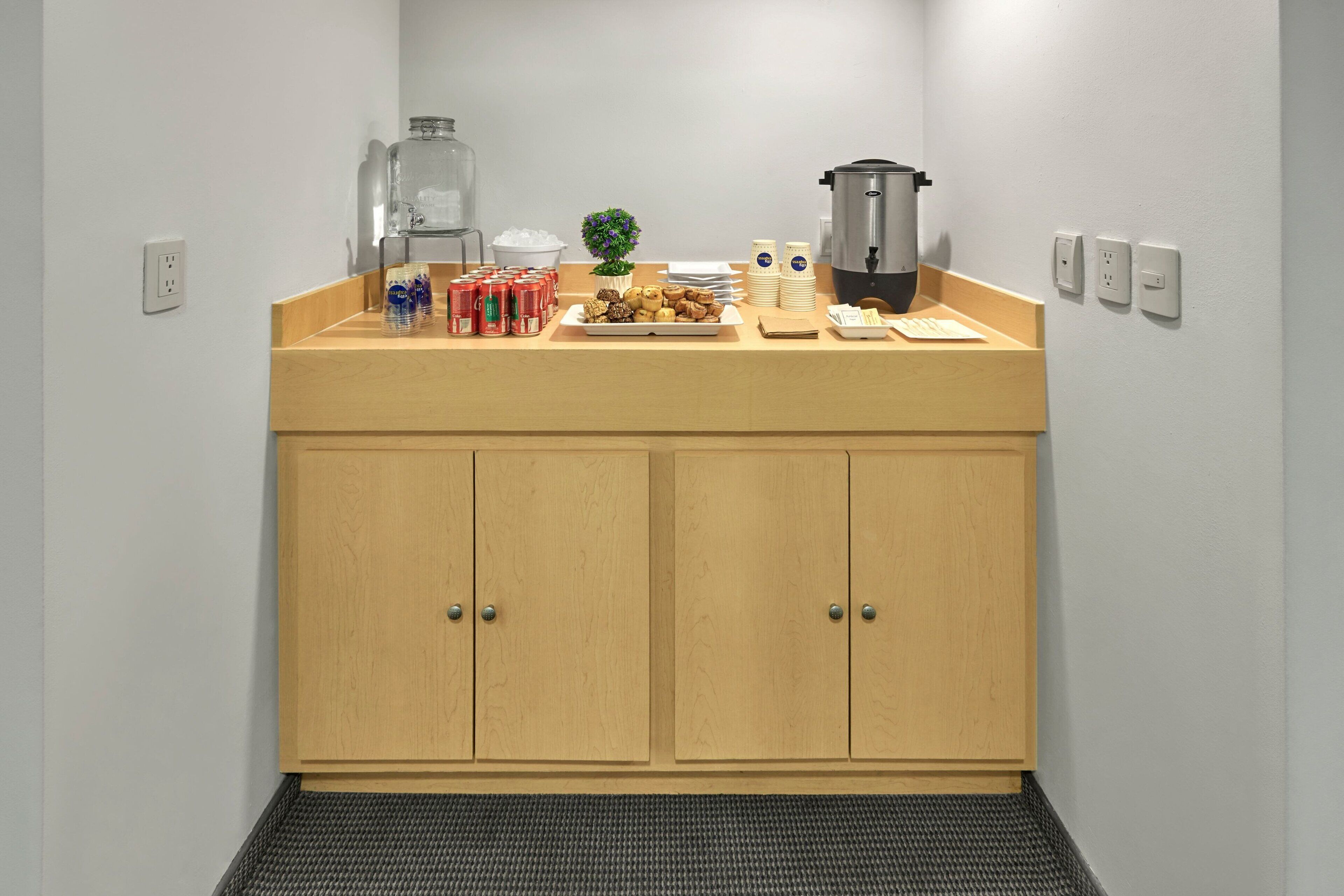 A kitchenette with a wooden countertop, a coffee maker, a kettle, and a small plant. There are also several cans and a plate of food.