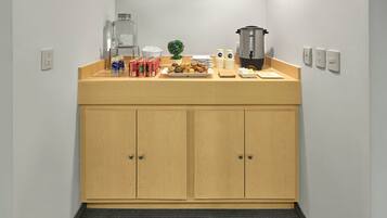 A kitchenette with a wooden countertop, a coffee maker, a kettle, and a small plant. There are also several cans and a plate of food.