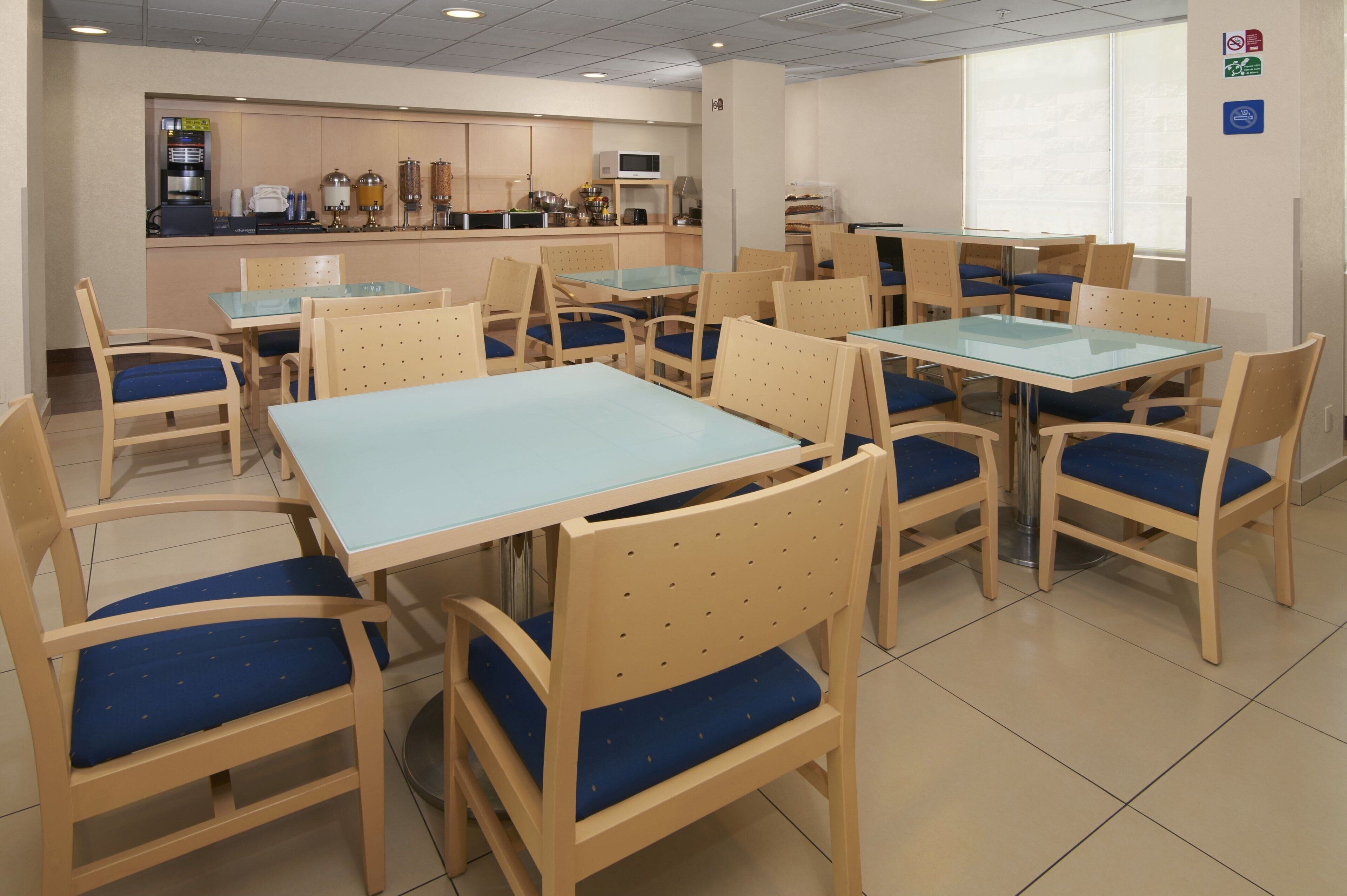 A dining area with tables and chairs, a kitchen in the background, and a window with blinds.
