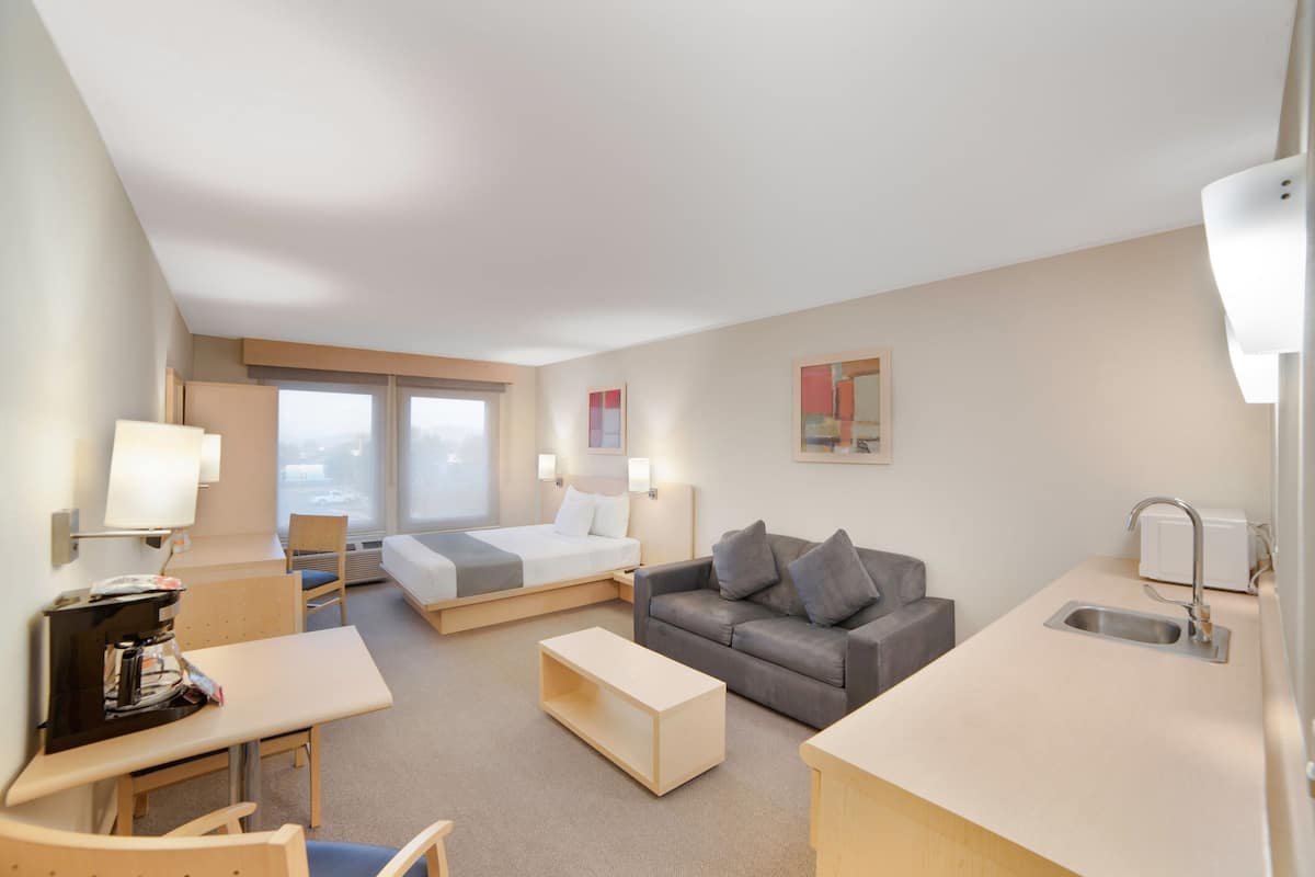 A hotel room with a bed, a sofa, a desk, a chair, a small kitchen area, and a window with a view.