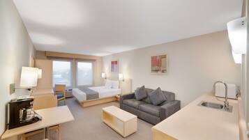A hotel room with a bed, a sofa, a desk, a chair, a small kitchen area, and a window with a view.