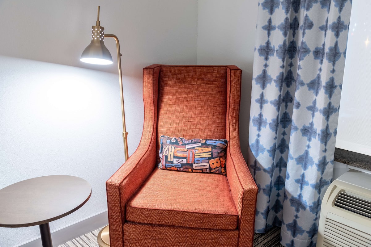 A room with a single orange armchair, a small round table, a floor lamp, and a patterned curtain.