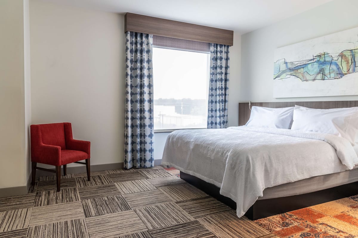 A hotel room with a large bed, a red chair, patterned carpet, and a window with curtains.